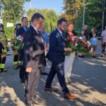 20240801_170318 Na obrazku grupa ludzi, w tym mężczyznach w garniturach i kobieta ubrana na biało, składane wieńce kwiatowe pod pomnikiem. Obok znajduje się strażacy i obserwatorzy kierujący ceremonii.