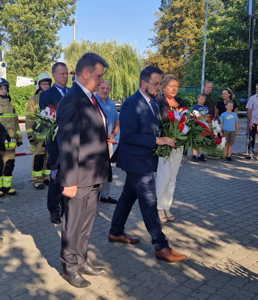 Na obrazku grupa ludzi, w tym mężczyznach w garniturach i kobieta ubrana na biało, składane wieńce kwiatowe pod pomnikiem. Obok znajduje się strażacy i obserwatorzy kierujący ceremonii.