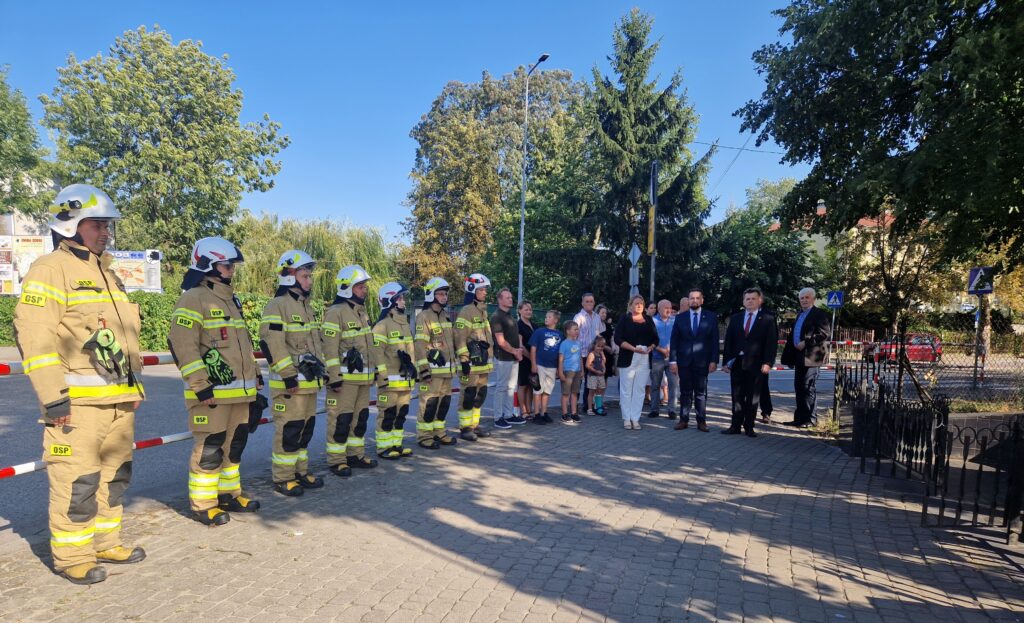 Na obrazku widać grupę izolatów stojących w szeregu na promieniach słonecznych, otoczoną przez ich ludzi, w tym ubranych w garnitury. W tle można używać drzewa.