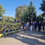 20240801_170408 Na obrazku widać grupę izolatów stojących w szeregu na promieniach słonecznych, otoczoną przez ich ludzi, w tym ubranych w garnitury. W tle można używać drzewa.