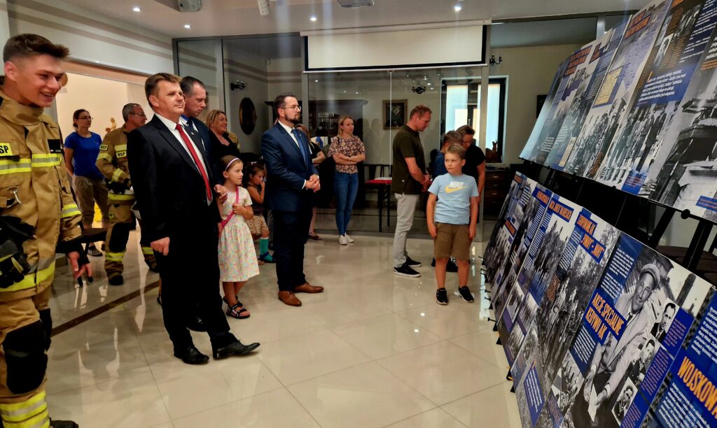 Grupa ludzi, w tym strażacy i dzieci, oglądają wystawę tradycyjnych informacji i fotografii na stojących panelach w eleganckim pomieszczeniu. Wszyscy mogą być zainteresowani prezentowanymi materiałami.