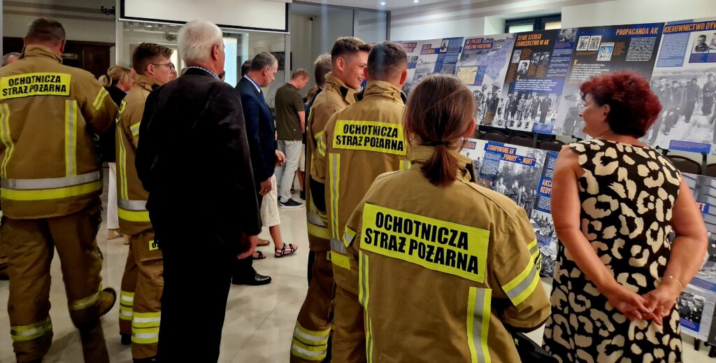 Na obrazku jest grupa ludzi, w tym strażacy ubrani w żółto-czarne uniformy z napisem „Ochotnicza Straż Pożarna”, którzy oglądają różne eksponaty w galerii. W tle widać kilka wystaw, przyciągających uwagę zwiedzających.