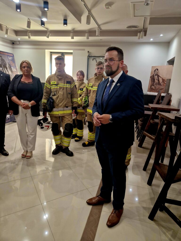 Na obrazku mężczyzny w garniturze mówić przed grupą osób, wśród których znajdują się strażacy w mundurach. Pokój jest jasnooświetlony i wyposażony w oprawę zdjęć na ścianach.