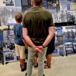 20240801_171555 Na obrazku trzy osoby blisko siebie, oglądając wystawę z historycznymi fotografiami i panelami tekstowymi. Cała scena ma miejsce w dobrze oświetlonym utworze.