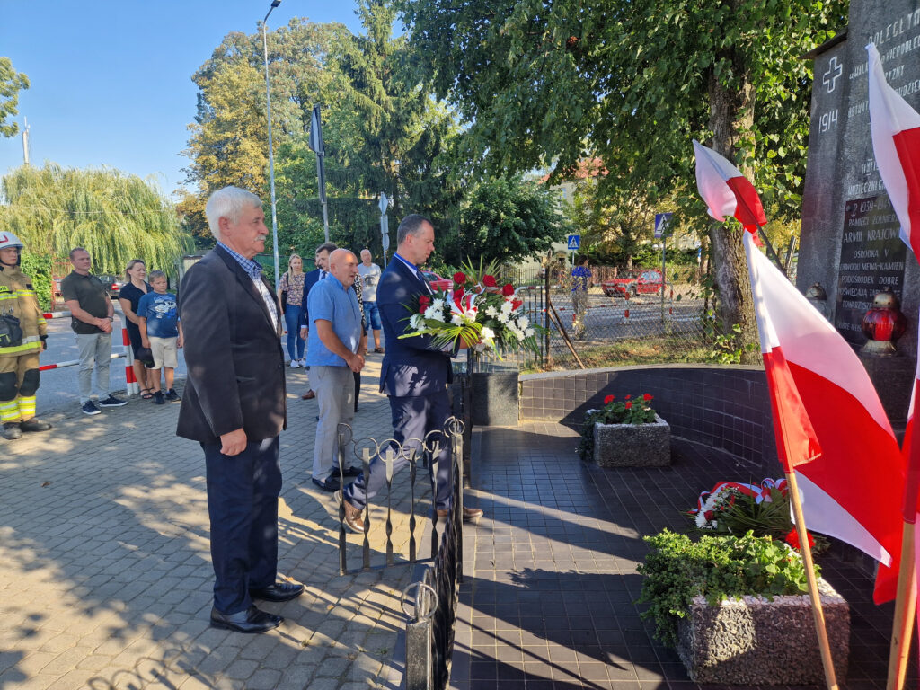 Na obrazku grupy mężczyzn, z których są ubrani w formalne stroje, umieszczając je na miejscu przy pomniku polskimi flagami. W tle znajdują się strażacy oraz osoby podlegające.