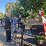 tinywow_20240801_170342_61483901 Na obrazku grupy mężczyzn, z których są ubrani w formalne stroje, umieszczając je na miejscu przy pomniku polskimi flagami. W tle znajdują się strażacy oraz osoby podlegające.