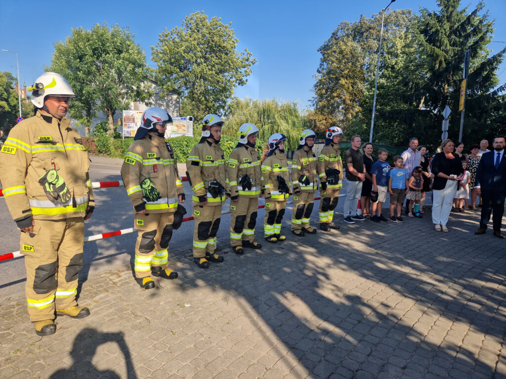 Na obrazie znajduje się grupa podlegająca w mundurach, ustalona na baczność przed tłumem obserwatorów, zarówno dzieci jak i dorośli. Cała scena ma miejsce podczas słonecznego dnia.