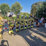 tinywow_20240801_170403_61483901 Na obrazie znajduje się grupa podlegająca w mundurach, ustalona na baczność przed tłumem obserwatorów, zarówno dzieci jak i dorośli. Cała scena ma miejsce podczas słonecznego dnia.