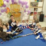 Na obrazie pojawia się grupa dzieci i dwóch dorosłych siedzących na niebieskiej macie w udekorowanej balonami i rysunkami z postaciami z kreskówek sali. Dzieci mają na sobie gotowe stroje, które są kompatybilne z niebieskimi tutu.