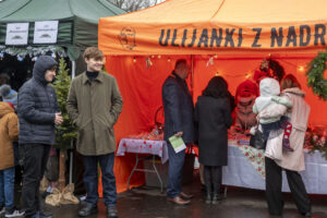 Na obrazku widać ludzi przeglądających stoiska na zewnętrznym rynku, udekorowanych świątecznymi targami. Pod namiotem wystawionym są towary, a jedna osoba trzyma dziecko, podczas gdy inni rozmawiają ze sprzedawcami.