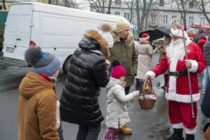 Na obrazku grupę ludzi ubranych w zimowe ubrania, którzy zgromadzili się wokół Świętego Mikołaja trzymającego kosz. Tło biały stanowi występny przy ulicy, co ważne, że scena ma miejsce w sezonie świątecznym.