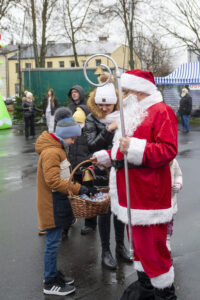 Na obrazku Świętego Mikołaja, który rozdaje smakołyki z koszyka dzieciom w świątecznej scenerii plenerowej. Wokół panuje radosna atmosfera, a uśmiechnięte dzieci cieszą się z prezentów.