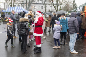 Na ekranie pojawia się grupa ludzi ubranych w zimowe stroje, przy czym osoba przebrana za Świętego Mikołaja wchodzi w interakcję z dziećmi. W tle są stoiska oraz drzewa, co charakterystyczne świąteczna atmosfera.