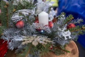 Na ekranie znajduje się świąteczna dekoracja stołu, której centralnym źródłem jest biała świeca otoczona czerwonymi ozdobami i srebrnymi gałązkami sosny. Elementy dekoracyjne obejmują figurkę renifera oraz napis „Wesołych Świąt”, przytulną, bożonarodzeniową atmosferę.