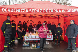 Na obrazku znajduje się grupa osób w mundurach stojących pod czerwonym namiotem z napisem „Ochotnicza Straż Pożarna”, a na stole przed nimi wystawionymi są różne przedmioty. Przed jednostką kobieta w różowym płaszczu mieszczącym się w jakimkolwiek obiekcie.