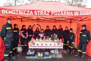 Na miejscu jest grupa kontrolna z Ochotniczej Straży Pożarnej w Dobrzycy, która jest pod czerwonym namiotem oznaczonym ich oznaczeniem. Przed nimi znajduje się krzesło, na którym znajdują się różne elementy, które mogą być powiązane z działalnością straży pożarnej.