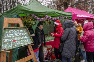 Na obrazku widać ludzi zgromadzonych przy udekorowanych stoiskach targowych, które pojawiają się świąteczne aranżacje i napisy. Atmosfera jest prawdopodobna radosna i przekazana z obchodami święta, jak Boże Narodzenie lub inny festiwal zimowy.