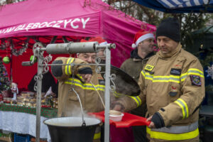 Na obrazie dwóch osobników w mundurach, którzy serwują jedzenie z garnka podczas plenerowej imprezy. W tle pojawia się różowe stoisko, co dodaje kolorystycznego akcentu do wydarzeń.