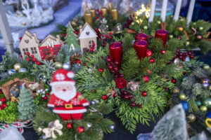 Na obrazie pojawia się wieniec bożonarodzeniowy, ozdobiony zielonymi gałązkami, czerwonymi świecami i jagodami. Widać figurkę Świętego Mikołaja, miniaturowe drewniane domki i różne dekoracje, przytulną świąteczną atmosferę.