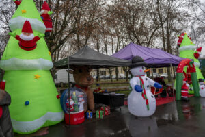 Na miejscu znajduje się świąteczna dekoracja dmuchana, obejmująca choinkę, bałwan i renifera pod namiotem na mokrym chodniku. W tle widać bezlistne drzewa.