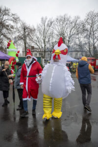 Na obrazku widać dwie osoby spacerujące na zewnątrz; jedna z nich jest przebrana za Świętego Mikołaja, a drugi kapelusz dziecka. Pogoda jest pochmurna, a chodnik się mokry.