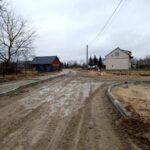 Na obrazku znajduje się błotnista droga w budowie, która może się znajdować na obszarach wiejskich z charakterystycznymi domami i drzewami. utrzymujący się także słupy energetyczne, a całość utrzymana jest w pochmurnej scenerii.