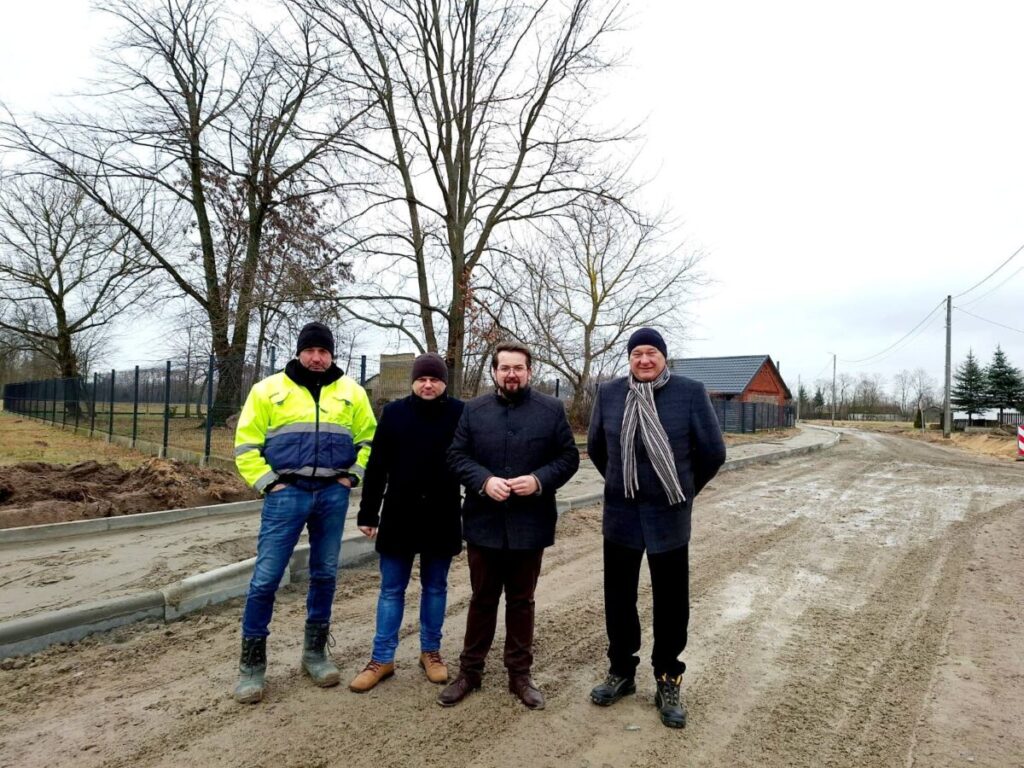 Na obrazku widoczne cztery osoby na błotnistej drodze w pochmurny dzień. W tle występuje drzewo oraz dom, tworzący wiejski, wiejski krajobraz.