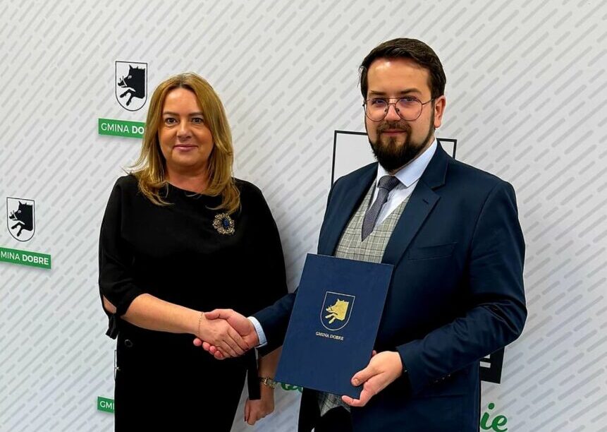 Na obrazku widać dwie osoby trzymające sobie dłonie; jedna z nich trzyma teczkę. Za nimi znajduje się tło z logotypami "Gmina Dobre".