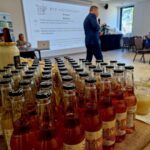 476606962_649843084057883_3441702295965295990_n Na pierwszym planie znajdują się butelki i pojemniki na stole. W tle widać osobę przemawiającą na terenie obok ekranu prezentacyjnego, a po prawej stronie siedziby.