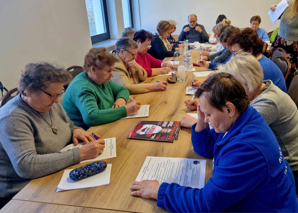 Na obrazku widać grupę osób siedzących wokół stołu, skupionych na pisaniu na kartkach papieru. Na miejscu znajduje się woda oraz widoczne są broszury lub ulotki.