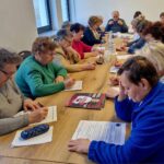 Na obrazku widać grupę osób siedzących wokół stołu, skupionych na pisaniu na kartkach papieru. Na miejscu znajduje się woda oraz widoczne są broszury lub ulotki.