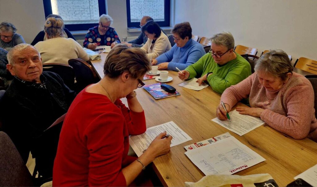 Starsza grupa osób siedzi przy drewnianym, dobrze oświetlonym pomieszczeniu, skupiając się na pisaniu i czytaniu dokumentów. Trzymają się w dłoniach długopisy, podczas gdy inni koncentrują się na zawartości przed sobą.