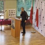 480480208_662748016100723_2483975698532487797_n Na obrazku widać mężczyznę przy mównicy, który mówi do mikrofonu w pomieszczeniu wyposażonym w flagami. Przed nim znajduje się stół z wyeksponowanymi różnorodnymi przedmiotami.