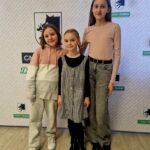 Na obrazku troje dzieci stoi przed tłem z logo i napisami, uśmiechając się do kamer. Dzieci wyglądają na zadowolone i są przeznaczone do walki z atakiem promocyjnym.