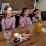 Na obrazku trójka dzieci siedzi przy stole, na którym znajdują się półmisek z wypiekami oraz butelka soku i herbaty. Dzieci są podatne na uszkodzenia w pomieszczeniach lub na drewnianych ścianach.