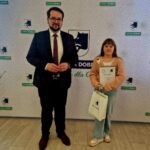 Mężczyzna i dziecko obok siebie na tle wzorzystej instalacji. Dziewczynka trzyma w ramionach dyplom oraz torebkę z prezentem.