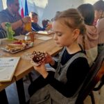 Na obrazku widać młodą dziewczynkę, która jest ozdobiona babeczkę, siedzącą przy ukryciu z inną osobą. Na ukrytym dyplomie znajduje się talerz z wypiekami.