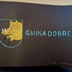 Na dole znajduje się umieszczona kartka, na której wyhaftowano żółtego dzika oraz napisy „GMINA DOBRE” na czarnym papierze. W osobnym dolnym rogu złotymi literami widnieje imię i nazwisko "LENA BARTNICKA".