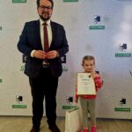 Na miejscu znajduje się dorosły i dziecko w pomieszczeniu; trzyma dyplom i prezenty na prezentach. W tle widoczny jest napis „Gmina Dobre”, co charakterystyczne, że scena ma miejsce podczas poszczególnych wydarzeń lub ceremonii wręczenia nagród.