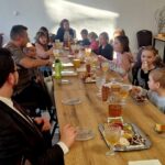 Na obrazku znajdują się grupy dorosłych i dzieci siedzących wokół długiego stołu, na których znajdują się produkty i wypieki. Spotkanie odbywa się w dobrze oświetlonym pomieszczeniu, co charakterystyczne i przyjazną atmosferę.