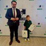 Na zdjęciu dorosły mężczyzna w garniturze obok dziecka, które trzyma w dłoni dyplom i miejsce zamieszkania. Obaj pozują do zdjęć na tle ściany z logo "Gmina Dobre".