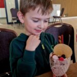 Na obrazku widać ubrane w bluzę z kapturem, które ujawniają dekoracyjne deserowi podanemu w szklance. Deser ozdobiony jest ciastkiem, waflowymi rurkami i jagodami, a całość znajduje się na drewnianym stole z wbudowanymi krzesłami w tle.