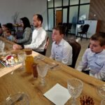 Na obrazku pojawia się grupa osób siedzących przy stole, który jest zastawiony napojami i przekąskami, w tym sokiem oraz półmiskami z deserami. Położenie wyróżniające się drewnianymi akcentami, a w tle są szklane drzwi.