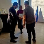 Na obrazku trzech ludzi stojących w pokoju, z których jedna osoba jest wręczona certyfikatowi drugiemu. W tle znajduje się stół z torbami prezentowymi oraz flaga Unii Europejskiej.