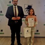 Na obrazku dorosła osoba w dodatku stoi obok dziecka, które trzyma certyfikaty oraz z prezentem. W tle jest dekoracja z herbem oraz napis „Gmina Dobre”, co oznacza oficjalna lub wręczenie nagród.