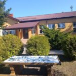 487219963_650510711174949_2720997157989791478_n Na zdjęciu widać żółty budynek z brązowym dachem, otoczony zielonymi krzewami. Na pierwszym planie stoi stół z mozaiką. W tle błękitne niebo i drzewa.