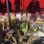 Na zdjęciu widoczne stoisko targowe z wbudowanymi produktami spożywczymi oraz wypiekami, a krzesło jest udekorowanym kwiatami i zielenią. Wokół ludzi w osłonach, znajdujących się pod czerwonym zadaszeniem.