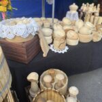 Na widocznym zdjęciu jest zestaw drewnianych przyborów kuchennych, takich jak miski, tłuczki i małe pojemniki, ułożone na czarnych obrusach wyposażonych w szydełkowanymi serwetkami. Dodatkowe dekoracje dopełniają kwiatowe akcenty związane między przedmiotami.