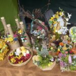 Na obrazku znajduje się aranżacja wielkiej nocy, w której znajdują się kolorowe pisanki, figurki króliczków, pierwotne kwiaty i wiklinowe koszyki na brązowym obrusie. Cała kompozycja emanuje świątecznym klimatem i wiosenną atmosferą.