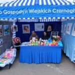 Na obrazku pojawia się niebiesko-biały pasiasty namiot, pod którym siedzą dwie kobiety za stołem, sprzedające kolorowe rękodzieła i przedmioty. Wokół namiotu wystawionego są różne oprawione dzieła sztuki.